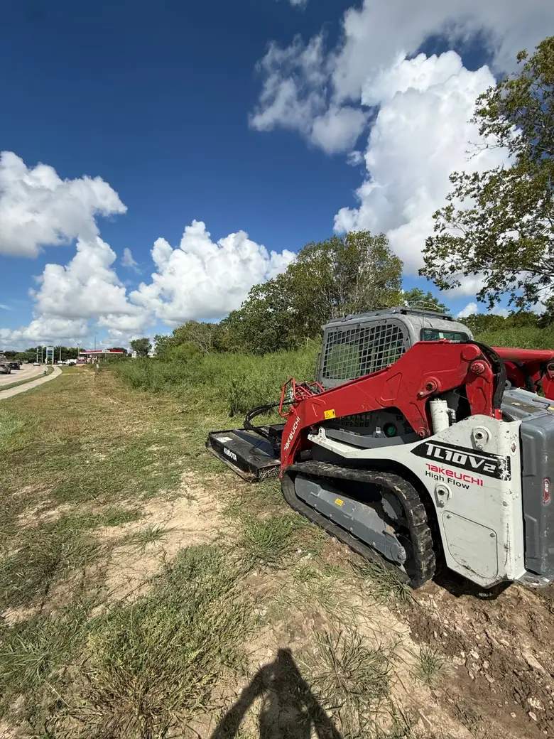 Takeuchi TL10V2 skid steer with a brush cutter attachment parked along the road frontage of the West Houston property.